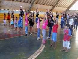Jovens e crianças fazem ginástica em quadra coberta.